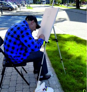 Mężczyzna maluje obraz na sztaludze, ulica, trawa, auta, chodnik